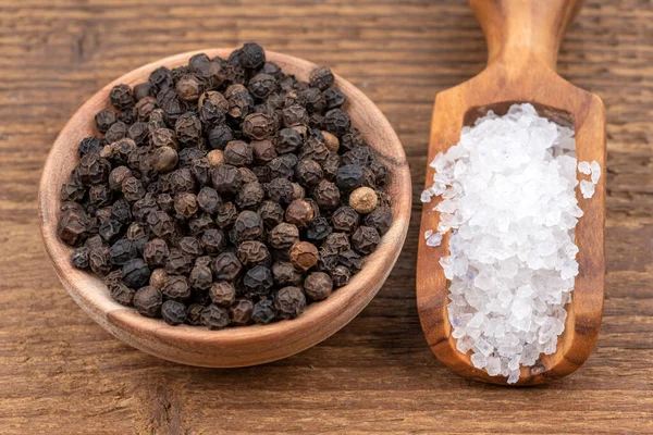 Coarse crystal salt on a small olive wood spice shovel and whole black peppercorns in a small wooden bowl on a rustic wooden background