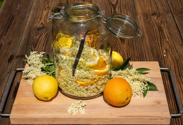 在切菜板上保存完好的大罐子里 放着新鲜的接骨木花 橙子和柠檬片 以及接骨木花甜酒用的香草豆荚 — 图库照片