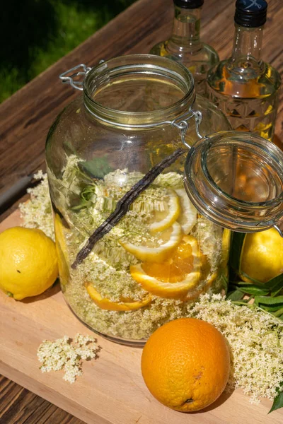 在切菜板上的大保鲜瓶里有新鲜的接骨木花 橙子和柠檬片 背景上的香草豆荚是接骨木花酒的伏特加瓶 — 图库照片