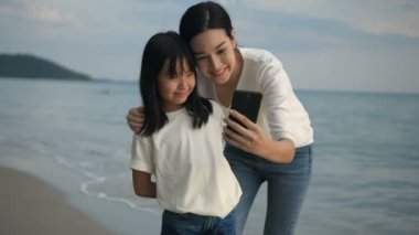Tatil konsepti. Anne ve kızı sahilde cep telefonlarıyla fotoğraf çekiyorlar. 4k Çözünürlüğü.