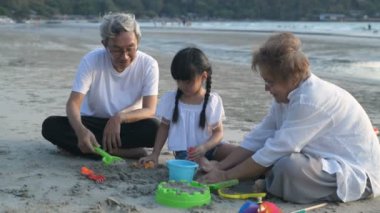 Tatil konsepti. Büyükbabam, büyükannem ve yeğenim kumsalda kum oynuyorlar. 4k Çözünürlüğü.