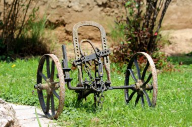 Eski, eski, paslı tarım ekipmanları metal çerçeveli ve aile evinin ön bahçesinde iki tekerlek bırakılmış bahçe süslemesi, ılık yaz gününde kesilmemiş çimenler ve bitkilerle çevrili.