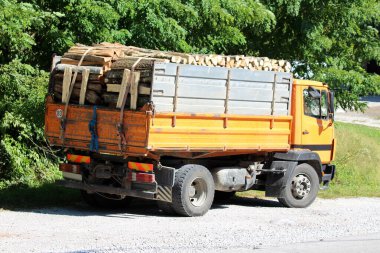 Sıcak yaz gününde, sık ağaçlarla çevrili asfalt yolun yanındaki çakıl parkına park edilmiş odunla dolu eski sarı kamyonun arka görüntüsü.