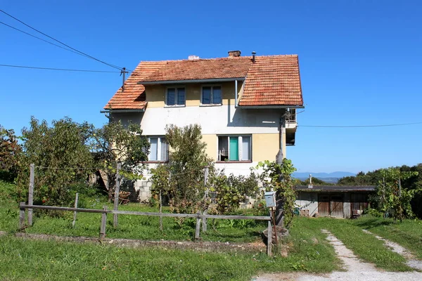 Tall suburban family house with dilapidated white yellow facade and faded color on roof tiles surrounded with uncut grass covered driveway and small trees on clear blue sky background on warm sunny summer day