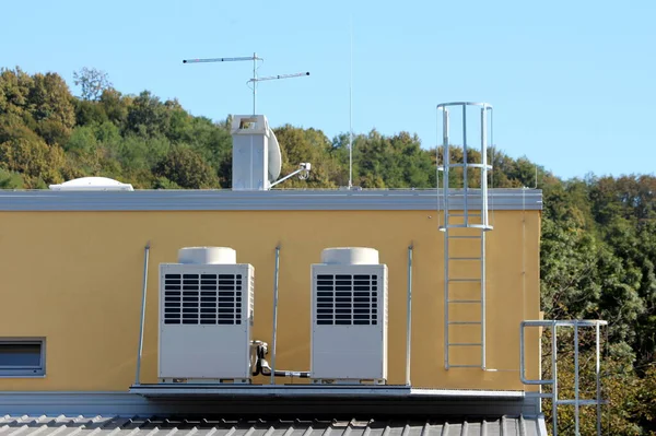 Two large ventilation units on metal platform on top of industrial building surrounded with metal stairs and dense forest in background on warm sunny summer day