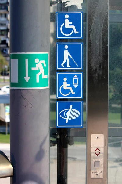Notification stickers with symbols for disabled persons and visually impaired persons on glass elevator entrance platform next to emergency exit symbol on warm sunny summer day