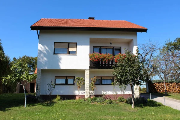 Beyaz banliyö aile evinin önü. Balkonunda kırmızı çiçekler ve yeni çatı fayansları. Kesilmemiş çimenler ve güneşli yaz gününde küçük ağaçlarla çevrili.