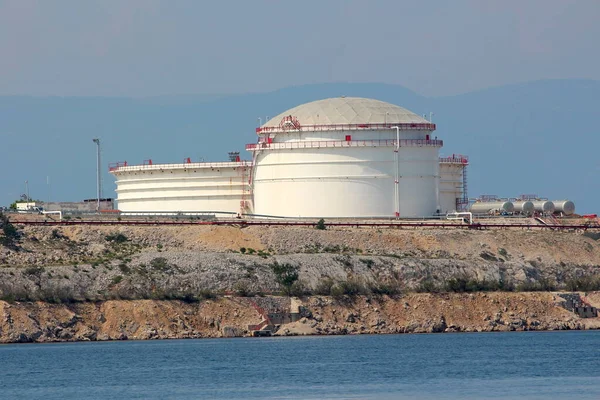 Large industrial white oil refinery tanks with protective fence placed on top of small hill surrounded with rocky shore and calm clear blue sea on warm sunny summer day