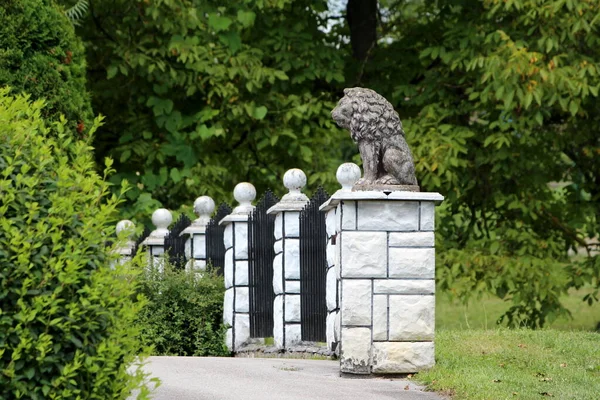 Family house driveway dark strong wrought iron fence mounted on white stone tiles covered poles with old lion sculpture on top surrounded with grass and dense trees on warm sunny summer day