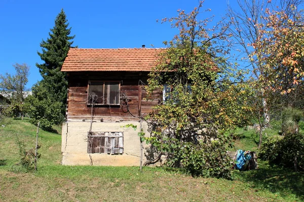 Dilapidated old wooden family house on top of tall concrete foundation with partially boarded window openings surrounded with freshly cut grass and trees on warm sunny summer day