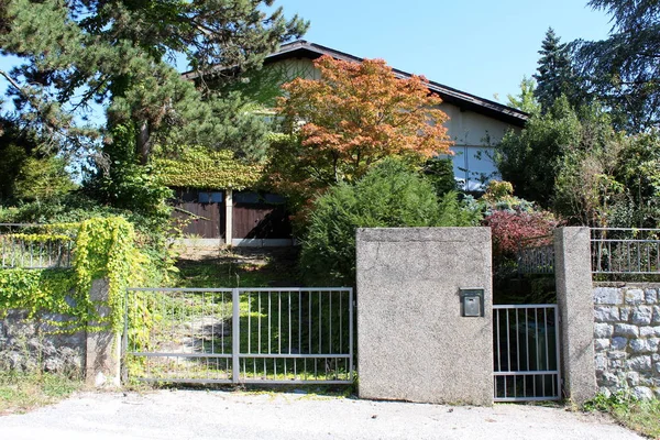 Suburban family house with overgrown driveway leading to two garage entrances almost completely hidden from public street with dense plants and trees surrounded with traditional stone wall and clear blue sky in background on warm sunny summer day