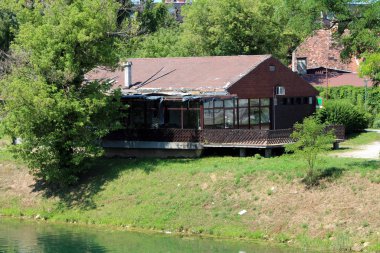 Abandoned small restaurant building with large front terrace and dilapidated destroyed roof built on local river bank surrounded with grass and dense trees