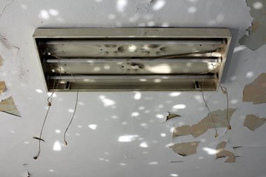 Broken overhead fluorescent lamp fixture with three fluorescent lamps and hanging disconnected electrical wires on cracked dilapidated ceiling at abandoned military complex on warm sunny summer day