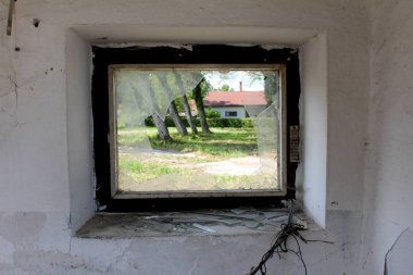 Broken small window with metal frame and shattered glass overlooking trees and buildings next to cut electrical wires on dilapidated white wall at abandoned military complex building on warm sunny summer day
