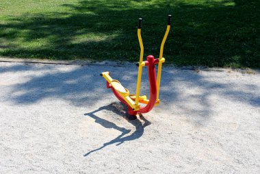 Eliptik çapraz antrenör. Renkli kırmızı ve sarı halka açık park spor ekipmanı. Çevresi çakıl ve yerel parktaki yeni biçilmiş çimlerle çevrili.