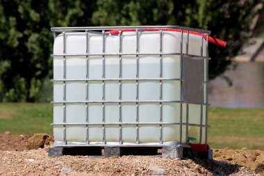 Su, arka planda sık ağaçlarla çevrili yerel inşaat sahasındaki çakıl yığınının tepesine metal kafesli IBC plastik tankı ya da orta büyüklükteki su dolu.