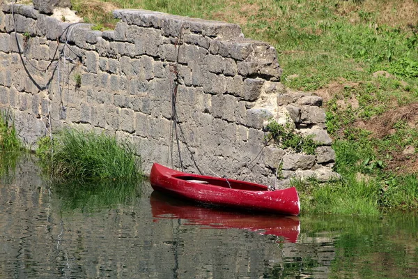 残破的深红色塑料独木舟绑在河岸的旧石墙上 周围环绕着平静的蓝河和青草 — 图库照片