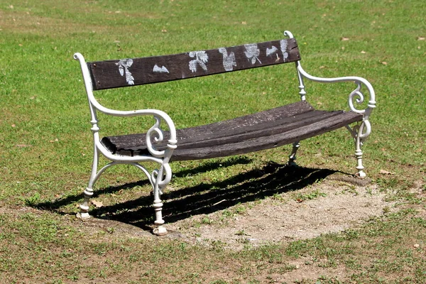 公園内には切妻草に囲まれた錬鉄製の支持体を持つバロック様式の木製のベンチの上面に白い色で描かれた紅葉 — ストック写真
