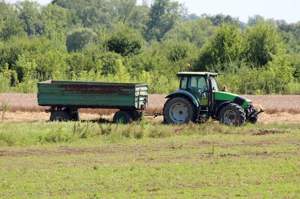 Green and black farm tractor with connected rusted vintage old tractor trailer left in local field surrounded with uncut grass and ripe wheat with dense forest vegetation in background