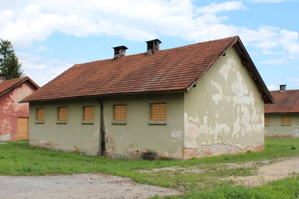 Green small dilapidated military barrack building with boarded windows and cracked facade surrounded with other buildings and uncut grass at abandoned military complex