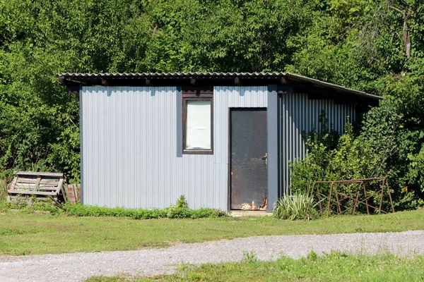 Homemade backyard tool shed storage area made from metal panels with doors and single window surrounded with grass gravel and dense forest vegetation in background