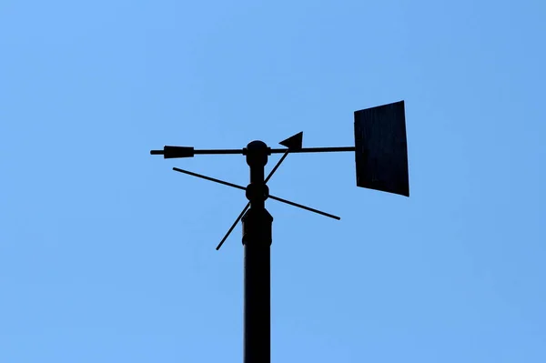 Silhouette of vintage retro weather vane instrument showing wind direction and arrow pointing towards north mounted on top of tall metal pole on clear blue sky background