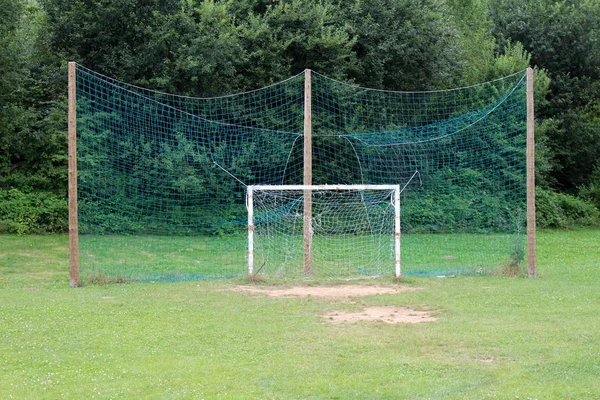 White dilapidated goal post for soccer practice with partially broken net in front of large green protection net suspended from tall wooden posts surrounded with grass and dense trees in background