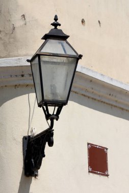 Eski barok tarzı sokak lambası. Siyah işlemeli demir kaplama, aile evinin yan tarafına monte edilmiş. Çatlak cephe, boş kahverengi sokak tabelasının yanında.