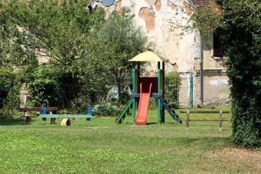 Tahterevalli şeklinde eski renkli açık hava oyun alanı ekipmanları ve merdivenlere tırmanan küçük ev ve arka planda umumi park bankları ve harap olmuş eski banliyö aile evleri ile çevrili kaydırak.