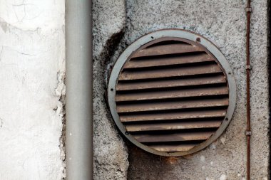 Paslanmış kısmen kapalı perdeleri olan yuvarlak havalandırma ızgarası harap olmuş aile evinin duvarına monte edilmiş yeni gri oluk borusunun yanında.