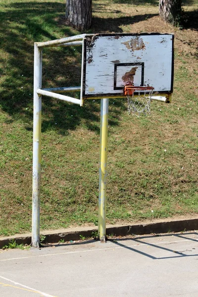 木製の板が割れた老朽化したバスケットボールのフープと 舗装されたバスケットボールコートと背景に新鮮な切口の草で囲まれた強い金属フレーム — ストック写真