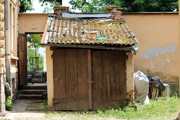 Garden tool shed with old wooden doors and dilapidated roof tiles covered with moss next to broken gutter in suburban family house backyard