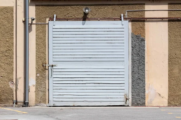 Heavy wooden and metal grey sliding storage doors mounted on dilapidated cracked wall of old fire station surrounded with gutter pipe and closed circuit TV CCTV security camera with paved parking lot in front and street lamp above