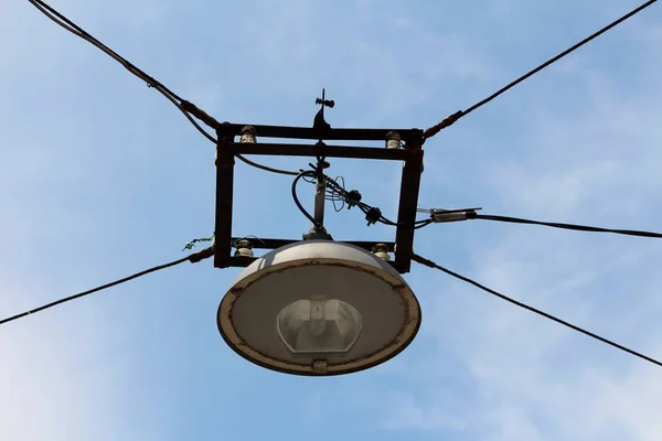 Large old street lamp within strong metal casing suspended with dark electrical wires in old part of town on cloudy blue sky background