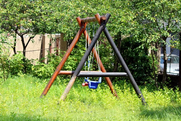 Massive old outdoor playground equipment swing with strong thick wooden frame and two plastic seats with faded colors surrounded with tall uncut grass and dense trees in suburban family house backyard