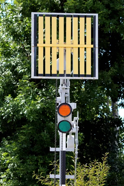 Railway trackside red and green traffic signal lights below signal sign mounted on metal pole at local train station with dense trees in background