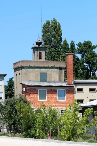 Red building blocks new unfinished part of old dilapidated fire station with vintage retro siren on top surrounded with gravel parking lot and tall trees in background