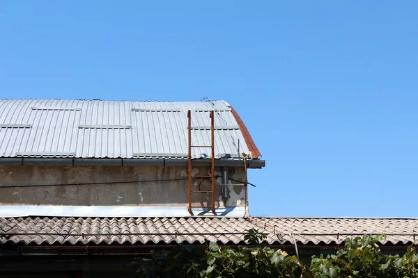 废弃的破旧不堪的工业综合体贮存库旁边的锈蚀金属梯子 上面覆盖着蓝天背景的金属屋顶瓦 — 图库照片