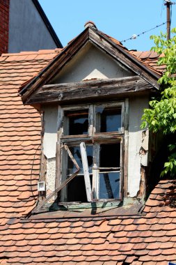 Kırık camlı ve kırık ahşap çerçeveli eski çatı penceresi terk edilmiş banliyö aile evinin tepesine monte edilmiş çatı kiremitleri ve küçük yapraklarla dolu yoğun ağaç dallarıyla çevrili.