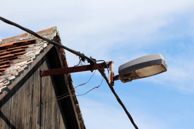 Paslı metal bir boruya monte edilmiş büyük kirli sokak lambası. Ahşap ahır çatısının kenarında, bulutlu mavi gökyüzü arka planında yeni koyu elektrik kabloları var.