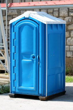 Yeni taşınabilir mavi ve beyaz ekolojik tuvalet ya da taşınabilir kimyasal tuvalet yerel inşaat alanındaki güçlü plastik temele monte edilmiş. Etrafı taşlarla kaplı banliyö aile evinin duvarlarıyla çevrili.