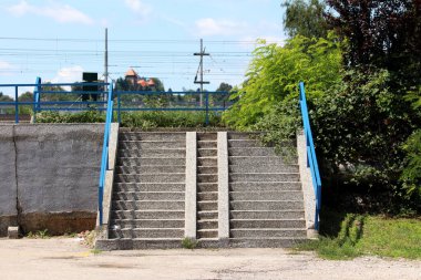 Arka planda yoğun santraller ve tren istasyonu kablolarıyla çevrili mavi tırabzan destekli eski beton halk merdivenleri.