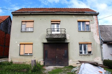 Eski bir banliyö aile evi harap olmuş ön cephesi ve diğer evlerle çevrili büyük ahşap çift giriş kapıları ve bulutlu mavi gökyüzü arka planında naylon korumayla kaplı odun yığını.