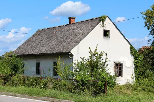 Abandoned small white suburban family house with broken windows and dilapidated facade covered with old roof tiles surrounded with overgrown tall uncut grass and various vegetation next to paved road on cloudy blue sky background
