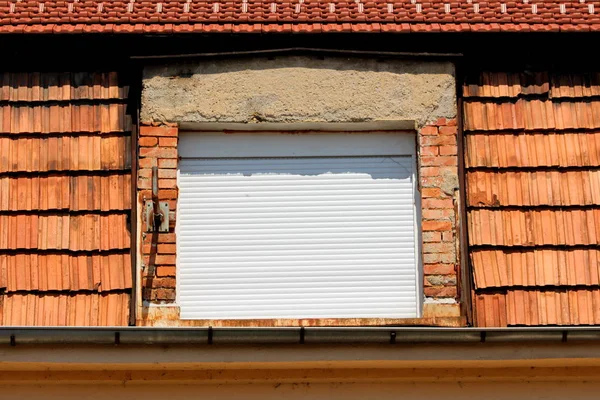 老朽化した割れ屋根瓦に囲まれた改装された屋根の下の郊外の家族の家の側に取り付けられた二重窓をカバーする新しいプラスチック製の窓ブラインドを閉じます — ストック写真