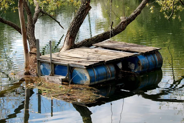 用蓝色金属桶和木板建成的自制简易河床固定在一棵大树上 周围是平静的河水和漂浮的青草 — 图库照片