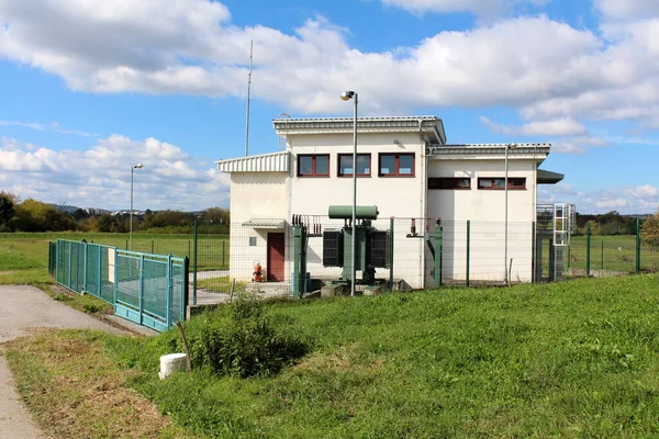 青い空を背景に 小さな窓と金属製の屋根がワイヤーフェンスと草で囲まれた地元の電気変電所の建物 — ストック写真