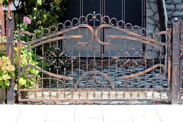 装飾的な要素を持つ巨大な強い錬鉄製のフェンスのドアは 花や庭の植物に囲まれた石のタイルで覆われた郊外の家族の家の私道へのアクセスを防ぐ — ストック写真