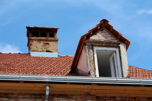 Old suburban family house under reconstruction with new roof window and roof tiles already installed surrounded with shiny new gutter next to cracked dilapidated red brick chimney and broken roof window wall on clear blue sky background