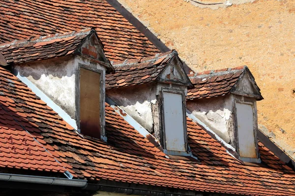 Row of old style roof windows closed with wooden boards covered with dilapidated red roof tiles and cracked facade on top of abandoned suburban family house in old part of town